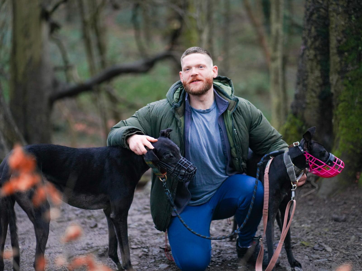 Garrett kneeling in the woods with Bran and Birdy, the two retired racing greyhounds behind Bran & Birdy’s
